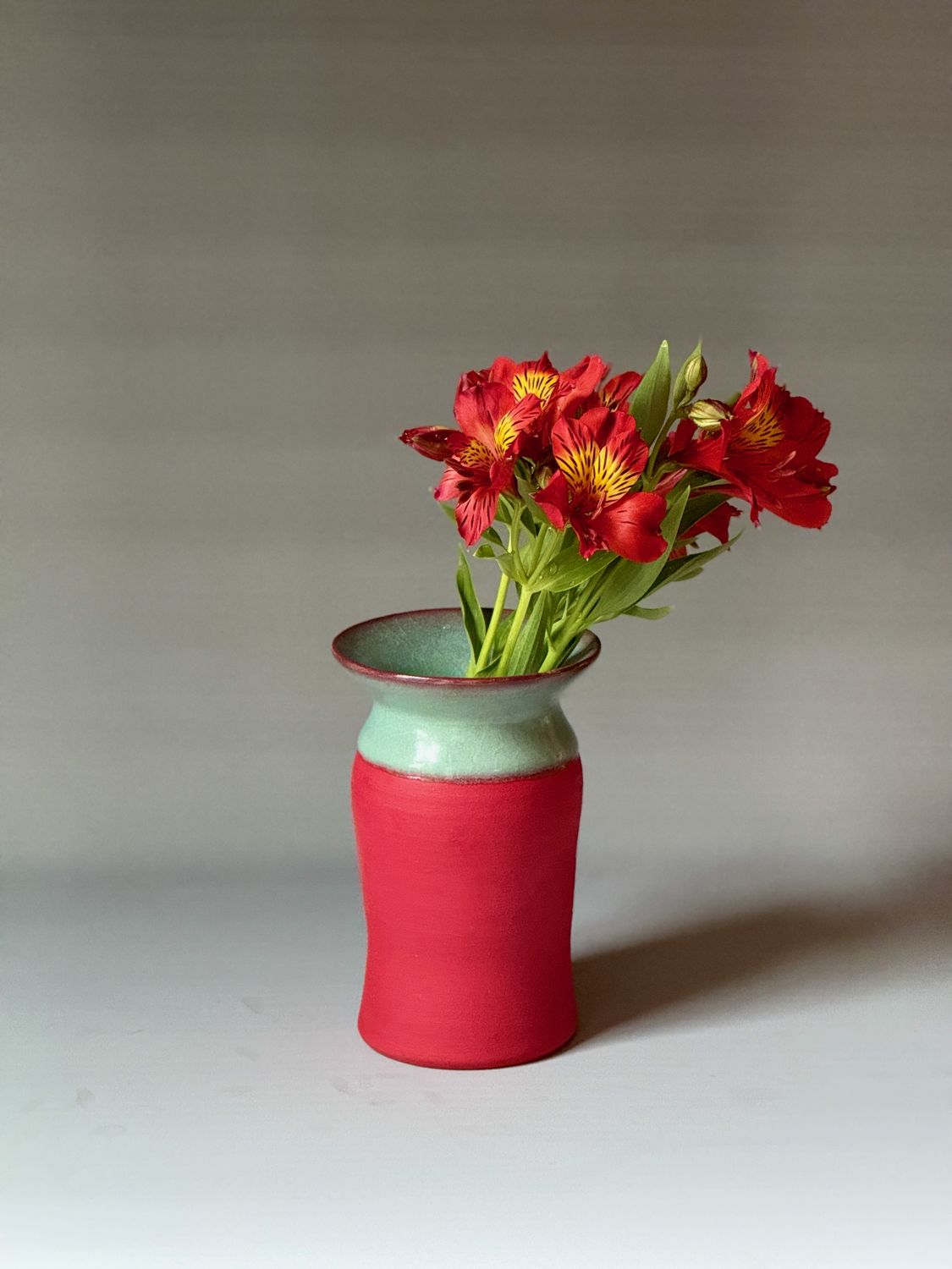 Poppy-red vase with a turquoise collar holding red alstroemeria, the vase from the logo